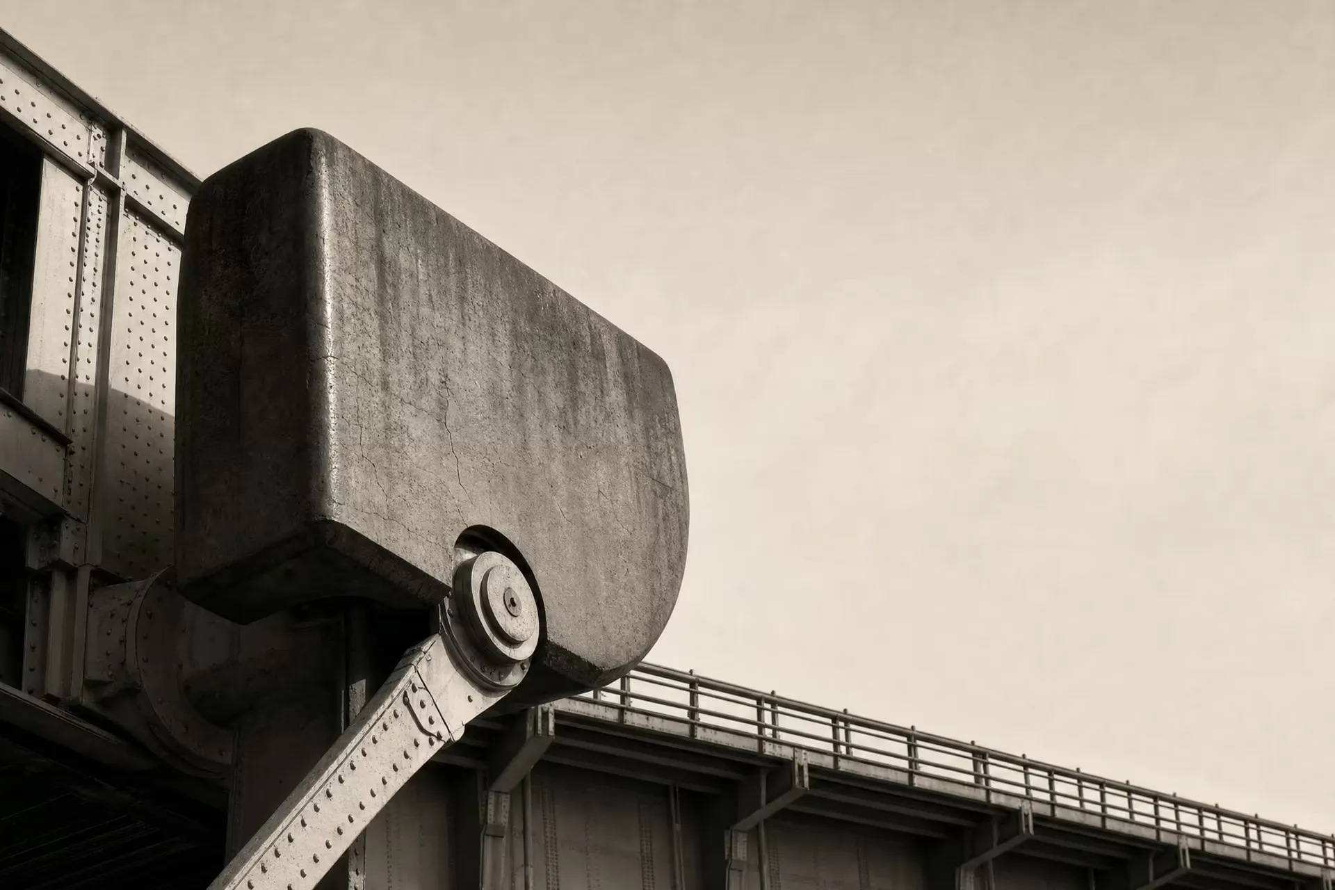 A massive bridge counterweight balanced on its pivot trunnion beneath the deck, riveted steel against open sky, black and white.