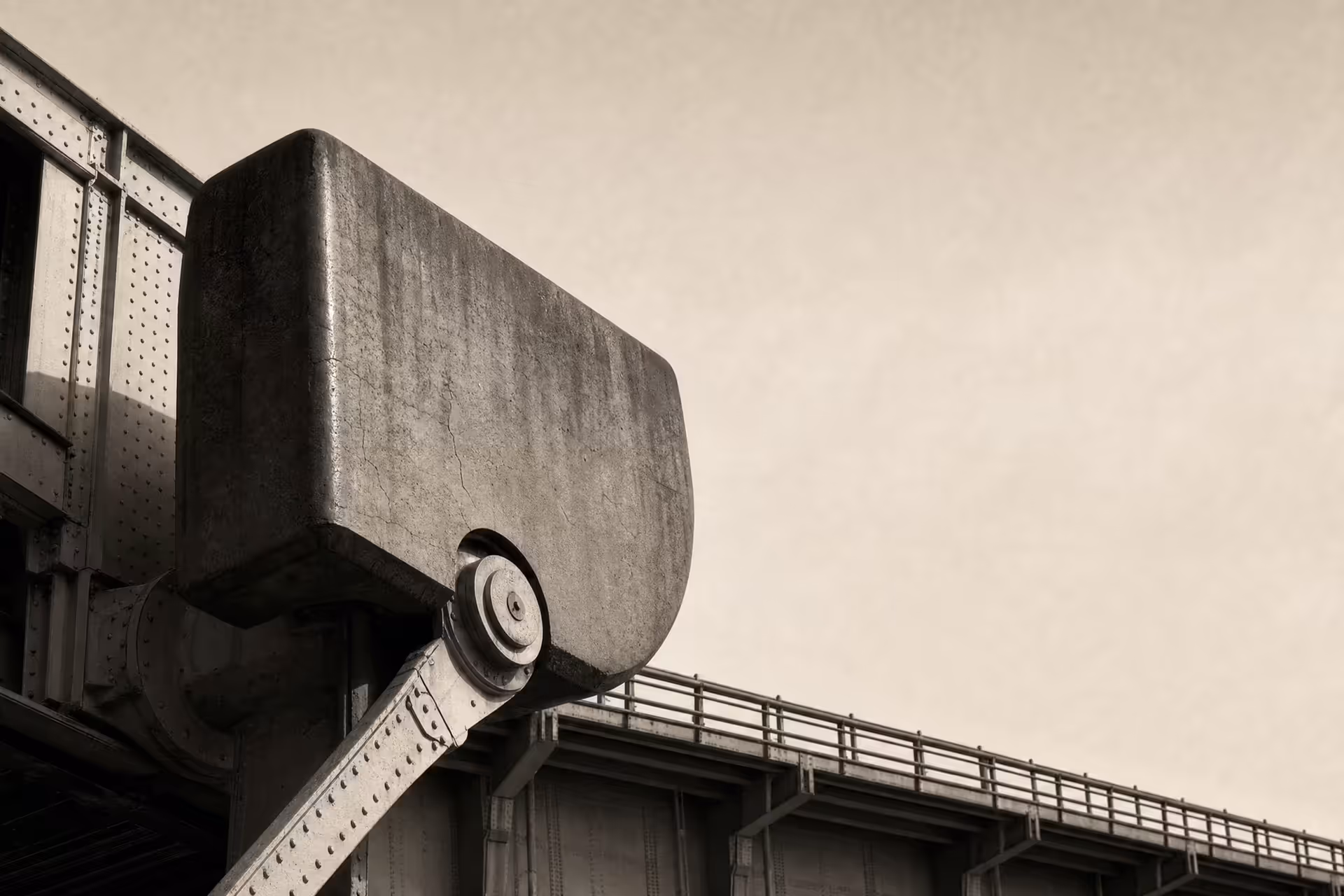 A massive bridge counterweight balanced on its pivot trunnion beneath the deck, riveted steel against open sky, black and white.