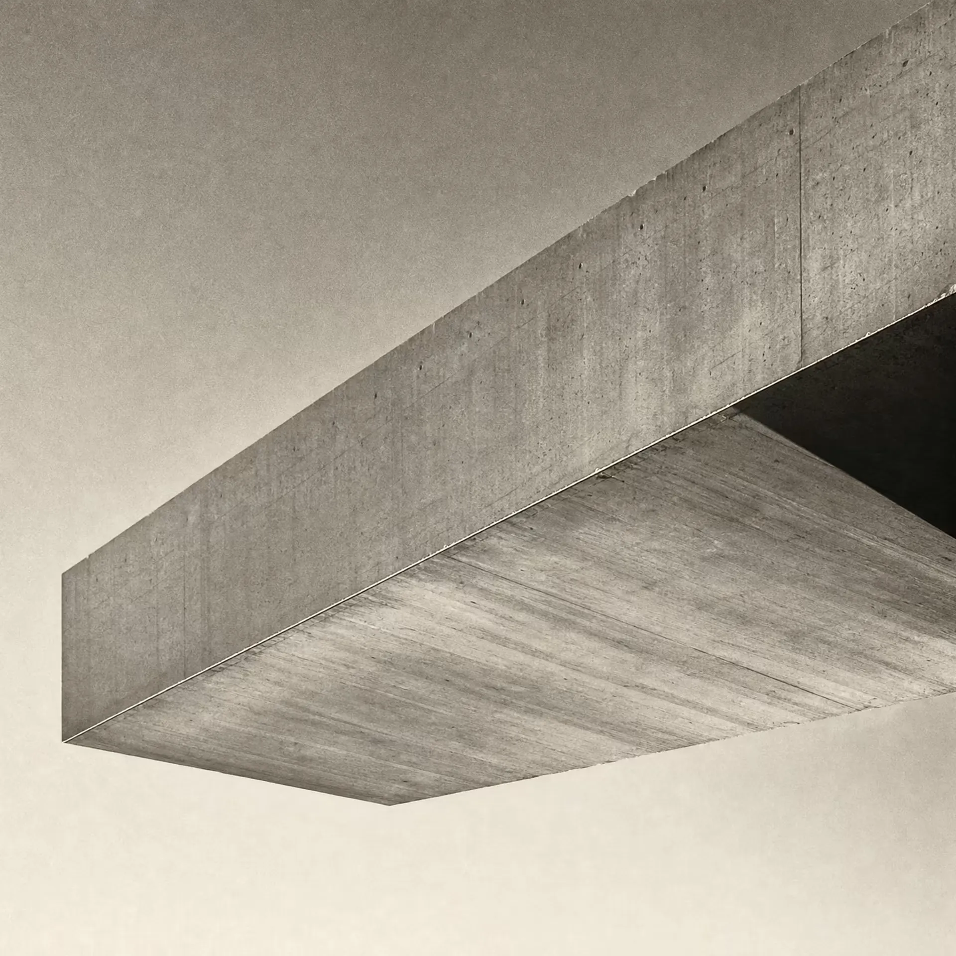A concrete cantilever beam projecting over open space from a single anchored support in warm-tone monochrome, the lit soffit reaching across an empty sky under grazing daylight.