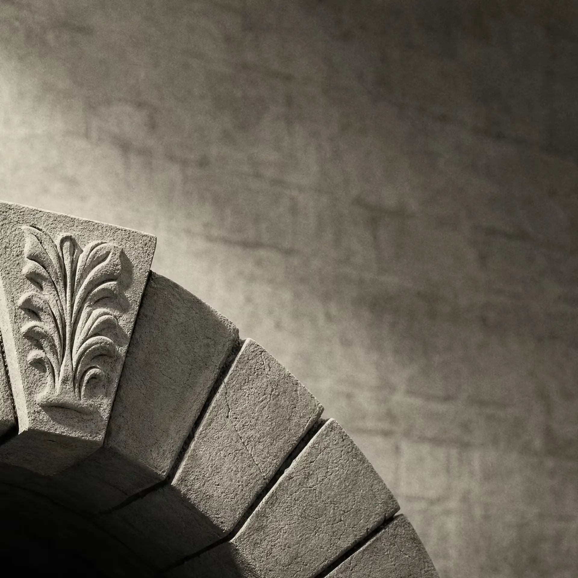 A single carved keystone locked at the apex of a stone arch in warm-tone monochrome, the wedge stones stepping down on either side under grazing light.