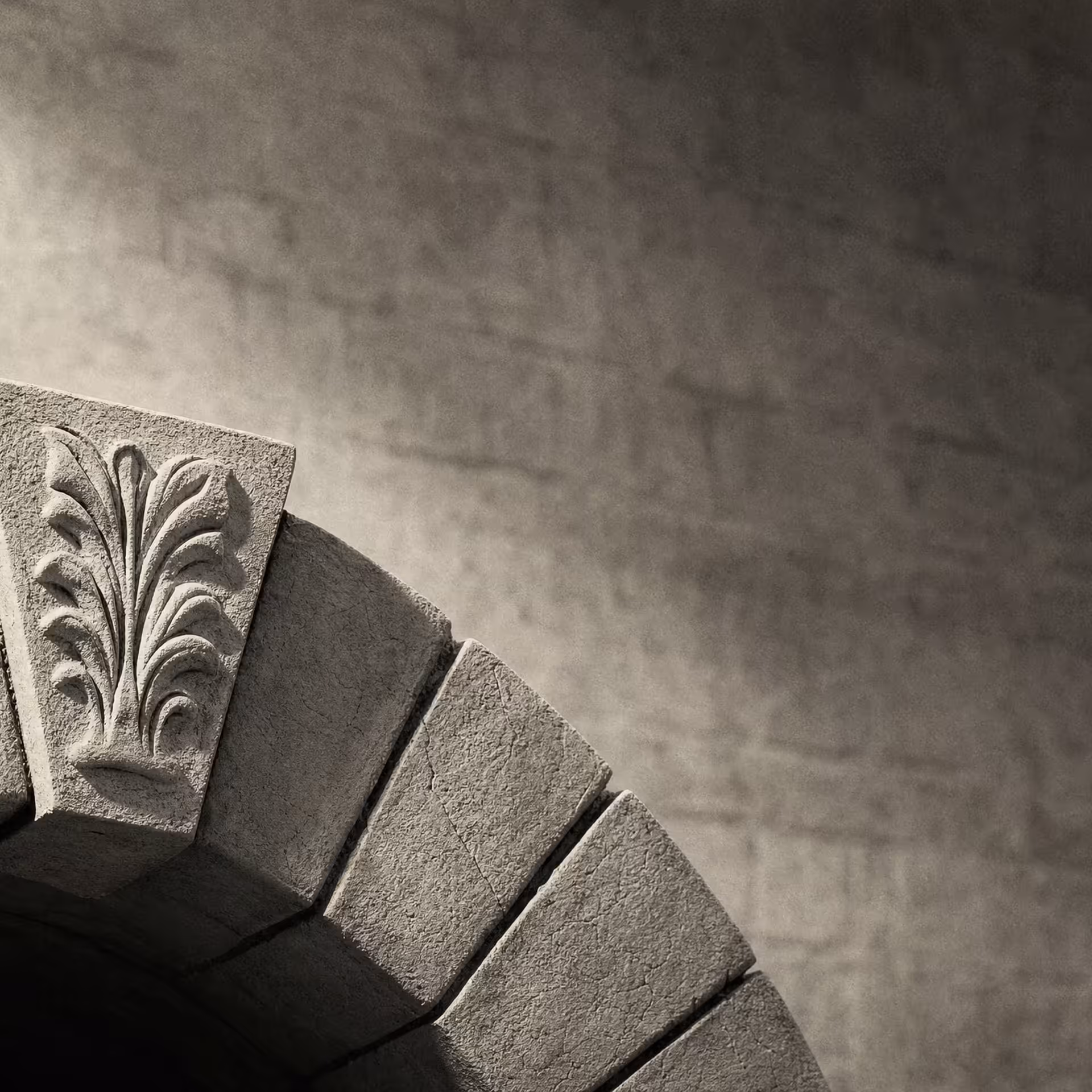 A single carved keystone locked at the apex of a stone arch in warm-tone monochrome, the wedge stones stepping down on either side under grazing light.