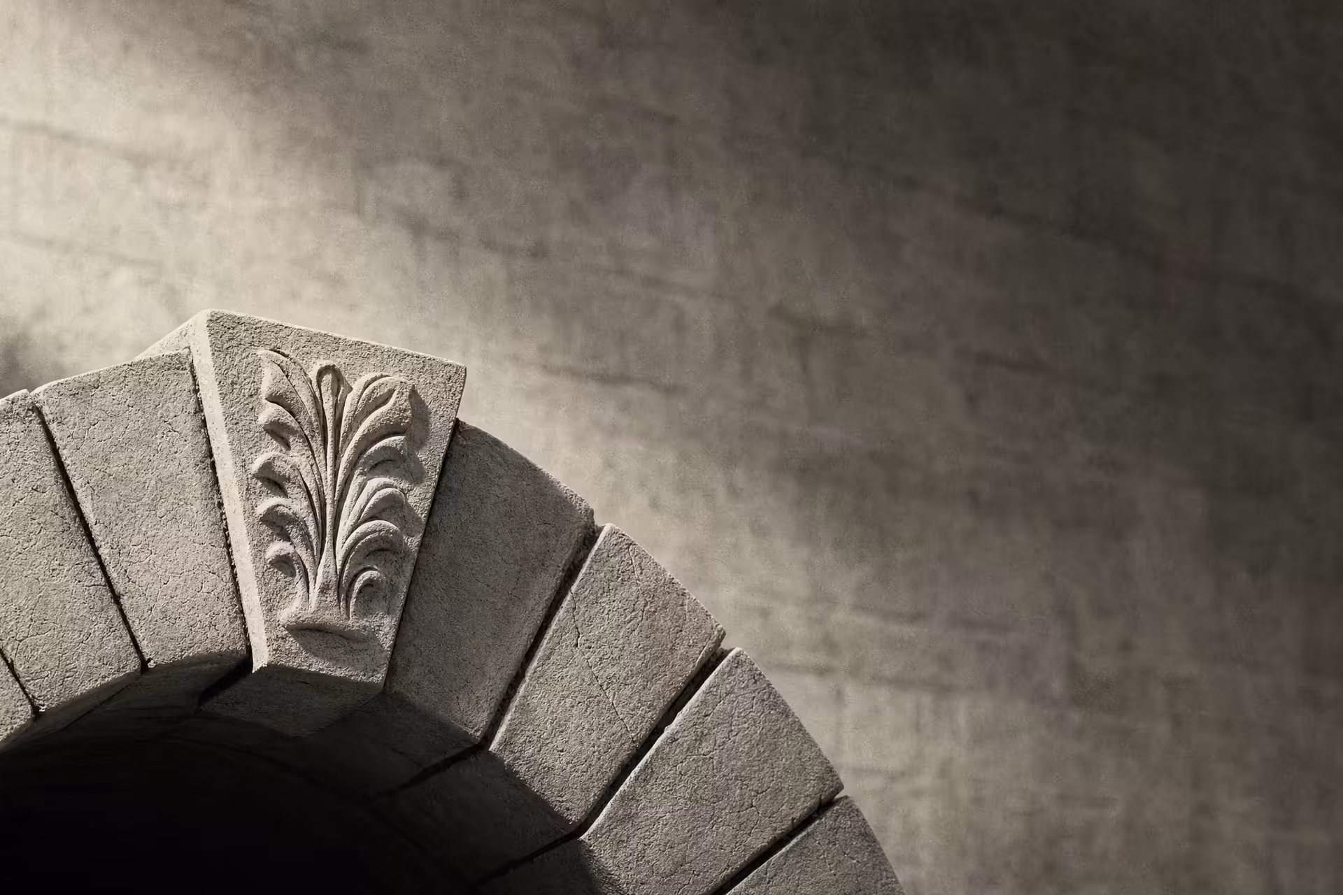 A single carved keystone locked at the apex of a stone arch in warm-tone monochrome, the wedge stones stepping down on either side under grazing light.