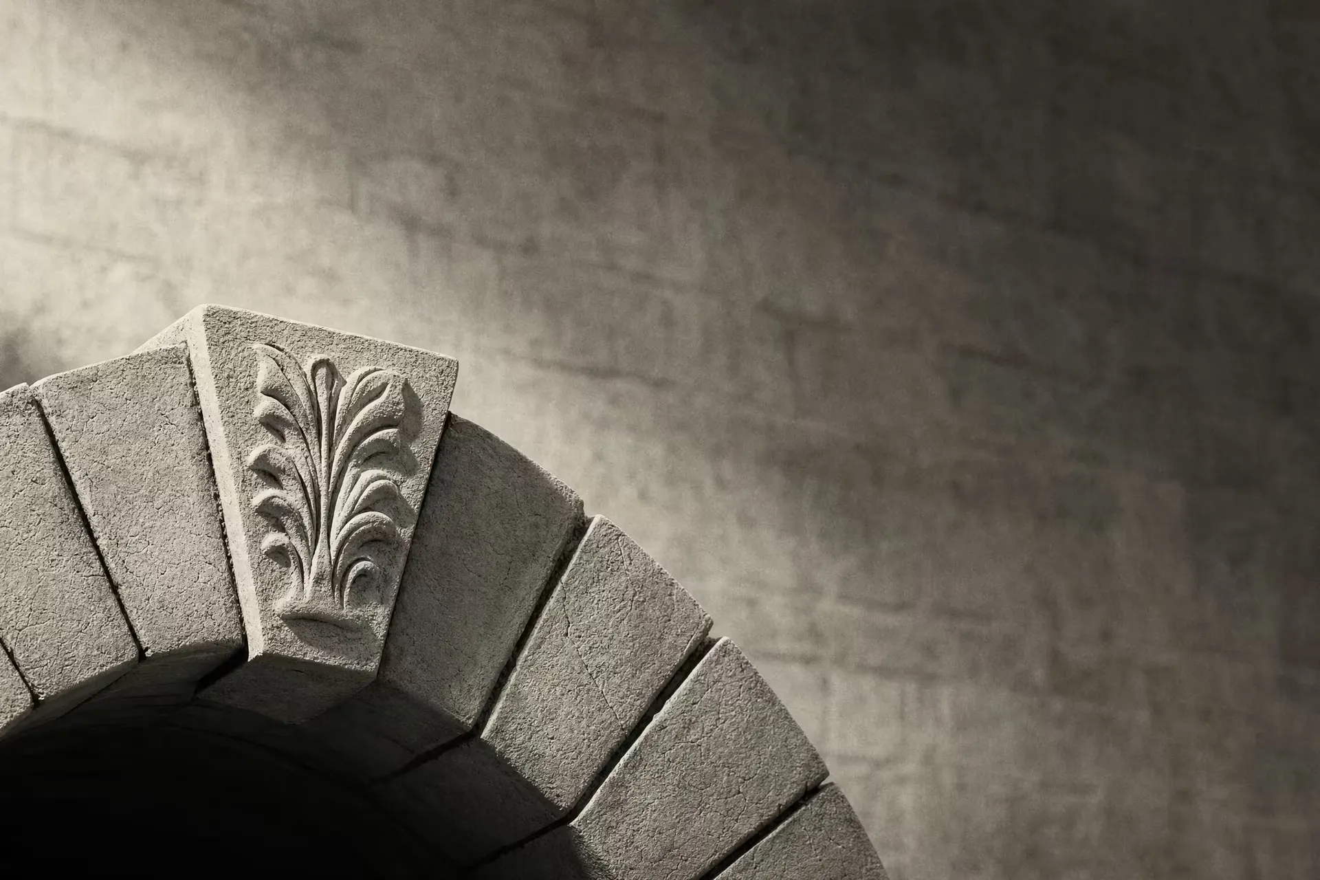 A single carved keystone locked at the apex of a stone arch in warm-tone monochrome, the wedge stones stepping down on either side under grazing light.