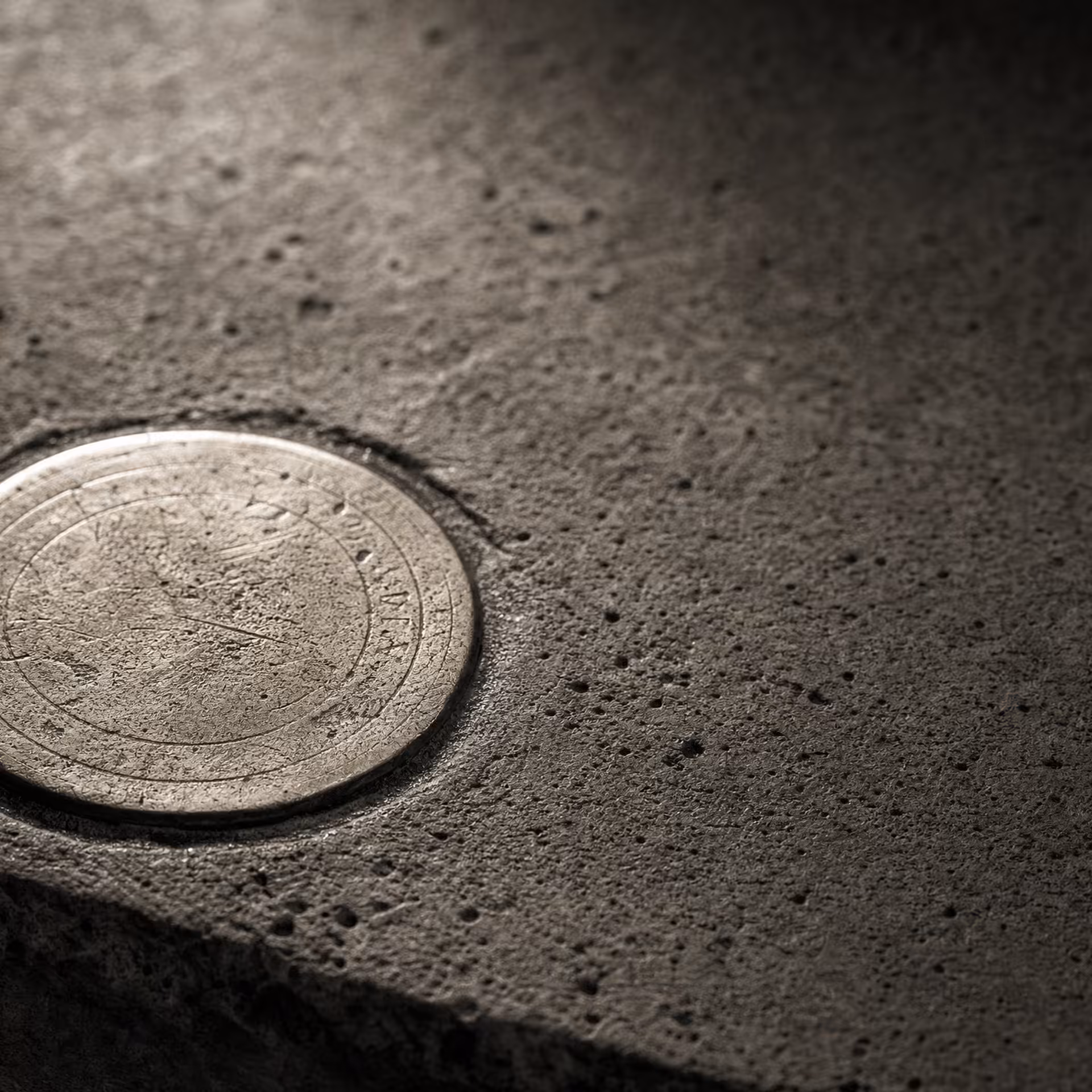 A circular brass survey datum disc set flush into weathered concrete in warm-tone monochrome, its worn reference rings catching a low raking light while the surrounding concrete grain falls into shadow.