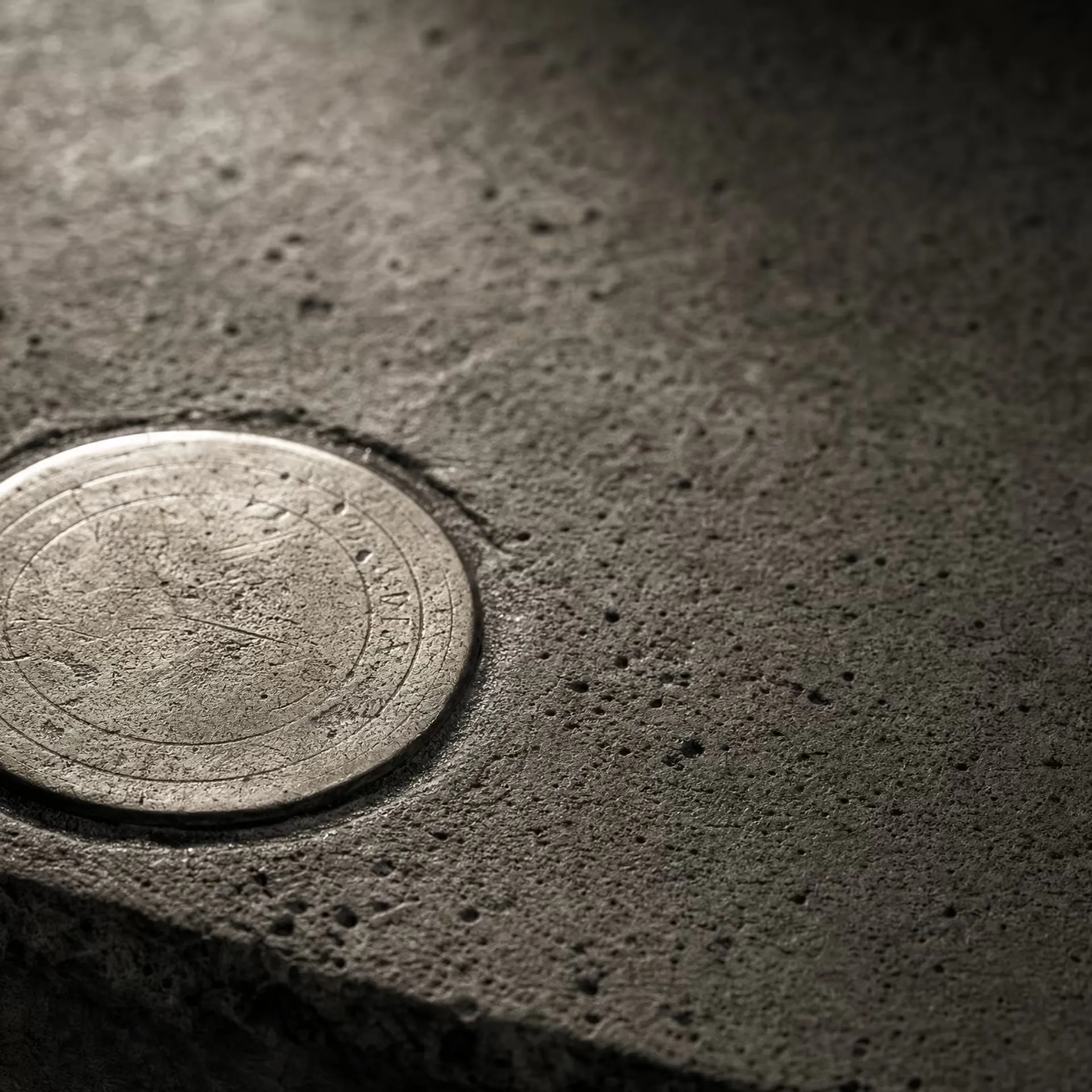 A circular brass survey datum disc set flush into weathered concrete in warm-tone monochrome, its worn reference rings catching a low raking light while the surrounding concrete grain falls into shadow.