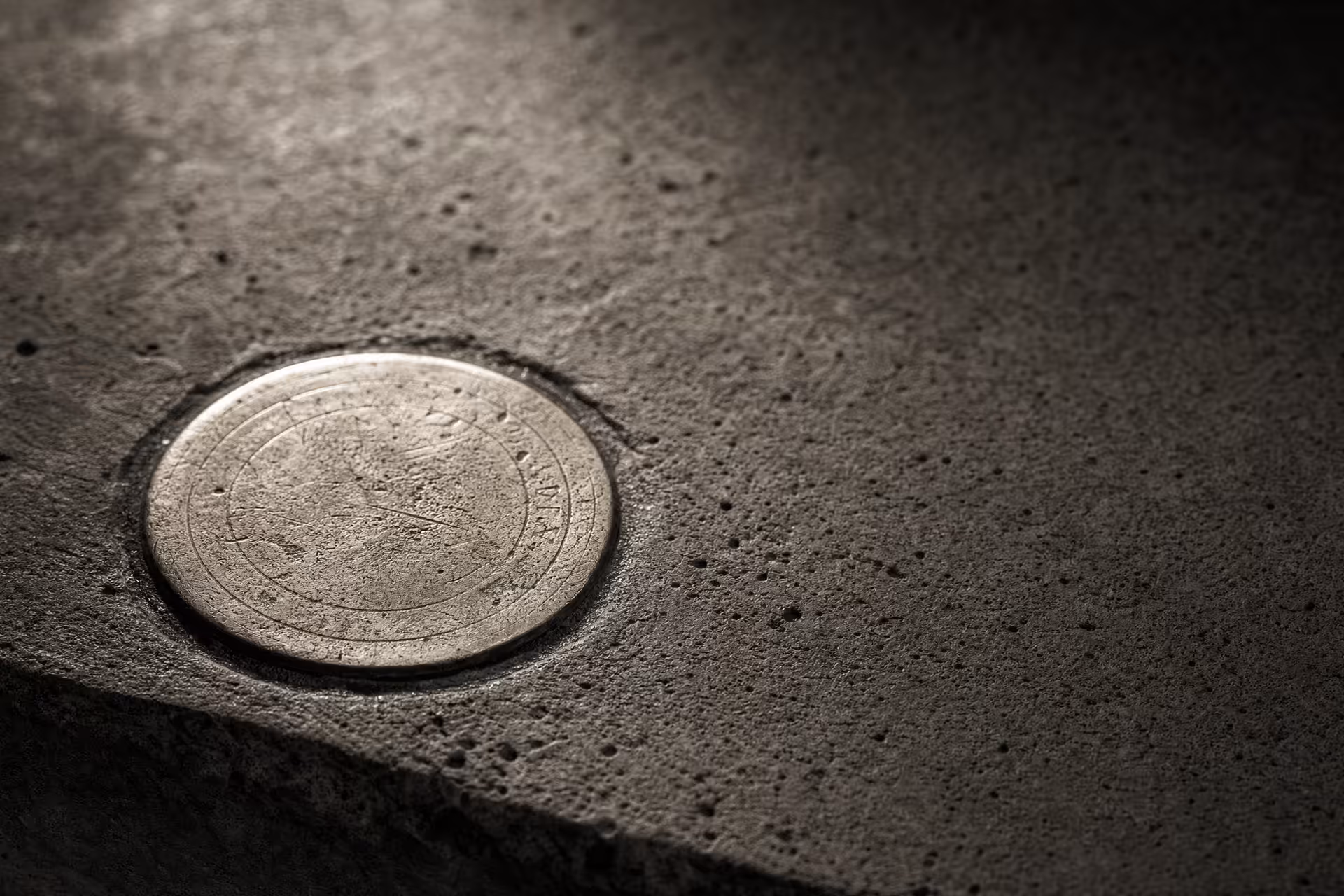 A circular brass survey datum disc set flush into weathered concrete in warm-tone monochrome, its worn reference rings catching a low raking light while the surrounding concrete grain falls into shadow.