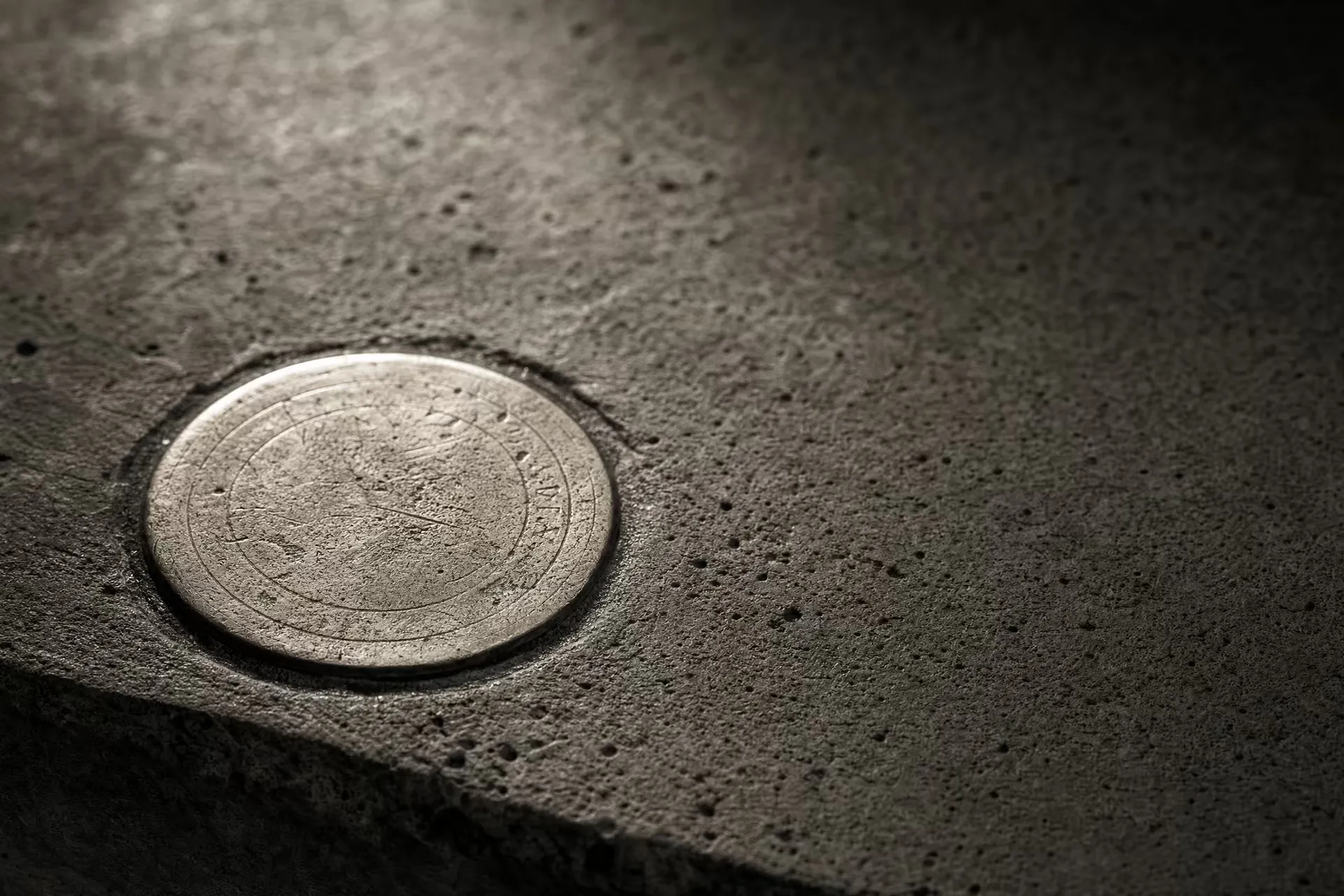 A circular brass survey datum disc set flush into weathered concrete in warm-tone monochrome, its worn reference rings catching a low raking light while the surrounding concrete grain falls into shadow.
