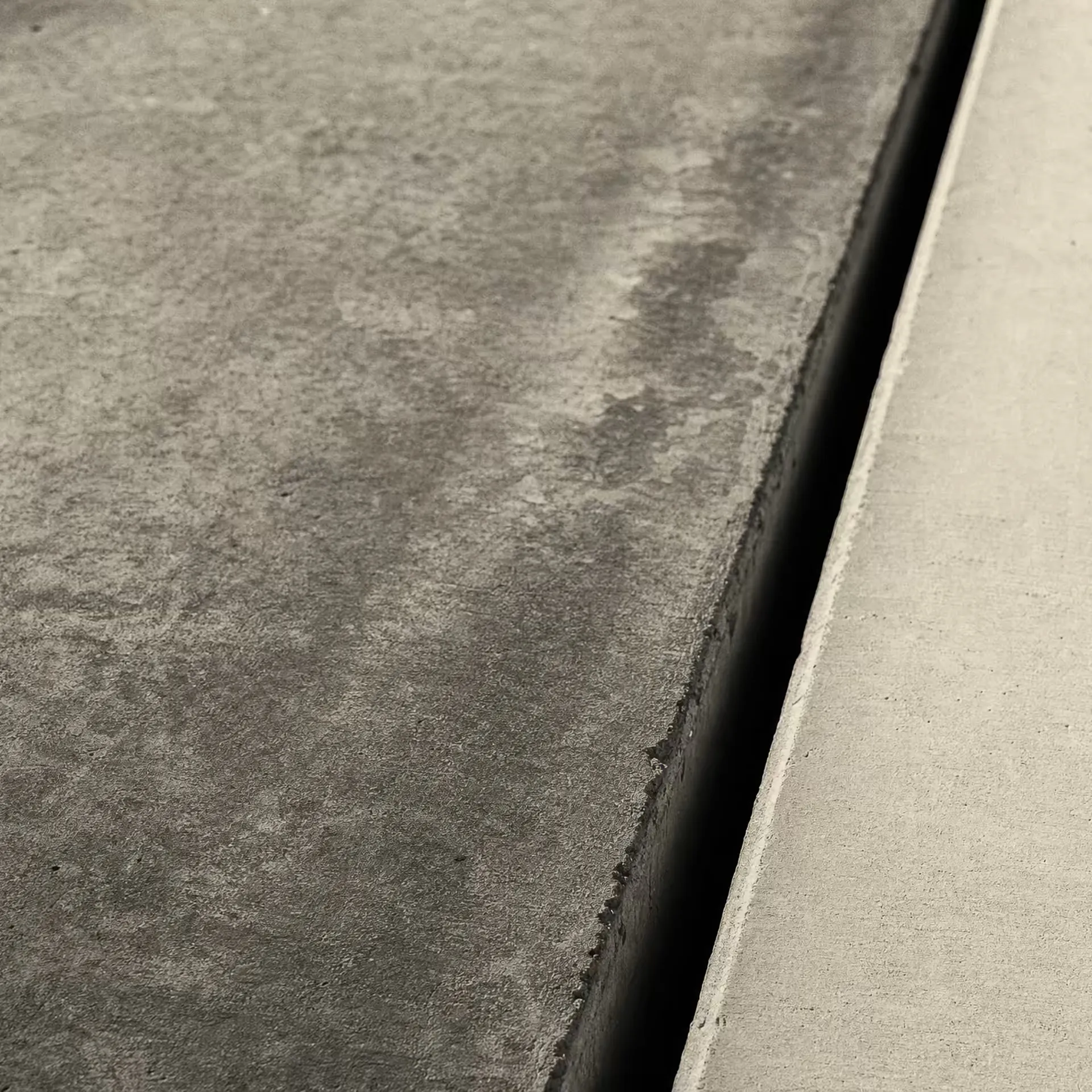 A concrete expansion joint in warm-tone monochrome, a dark vertical seam dividing an older weathered slab from a newer smooth one, a low raking light revealing the texture difference across the gap.