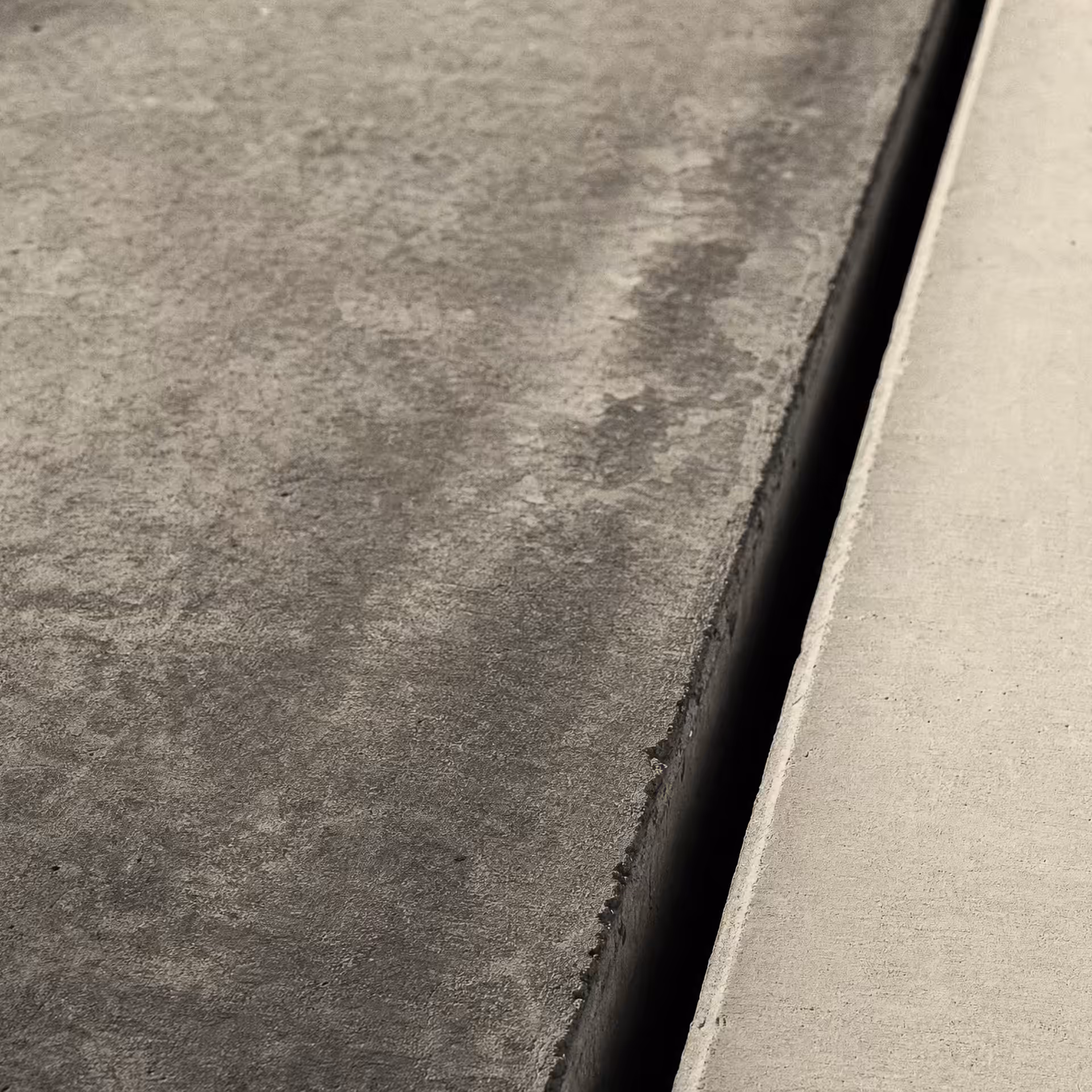 A concrete expansion joint in warm-tone monochrome, a dark vertical seam dividing an older weathered slab from a newer smooth one, a low raking light revealing the texture difference across the gap.
