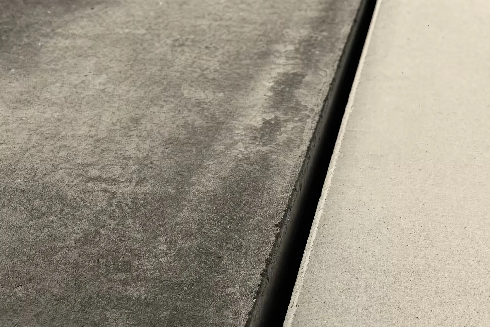 A concrete expansion joint in warm-tone monochrome, a dark vertical seam dividing an older weathered slab from a newer smooth one, a low raking light revealing the texture difference across the gap.