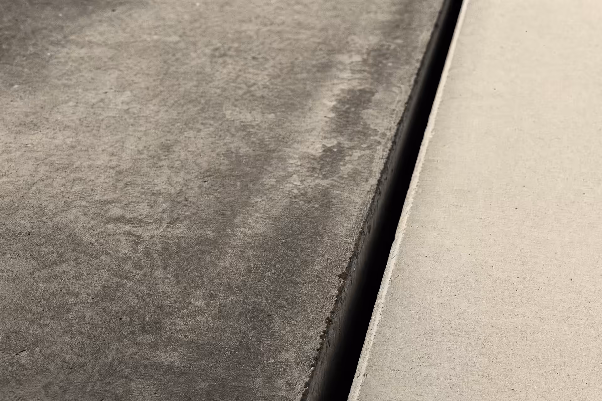 A concrete expansion joint in warm-tone monochrome, a dark vertical seam dividing an older weathered slab from a newer smooth one, a low raking light revealing the texture difference across the gap.