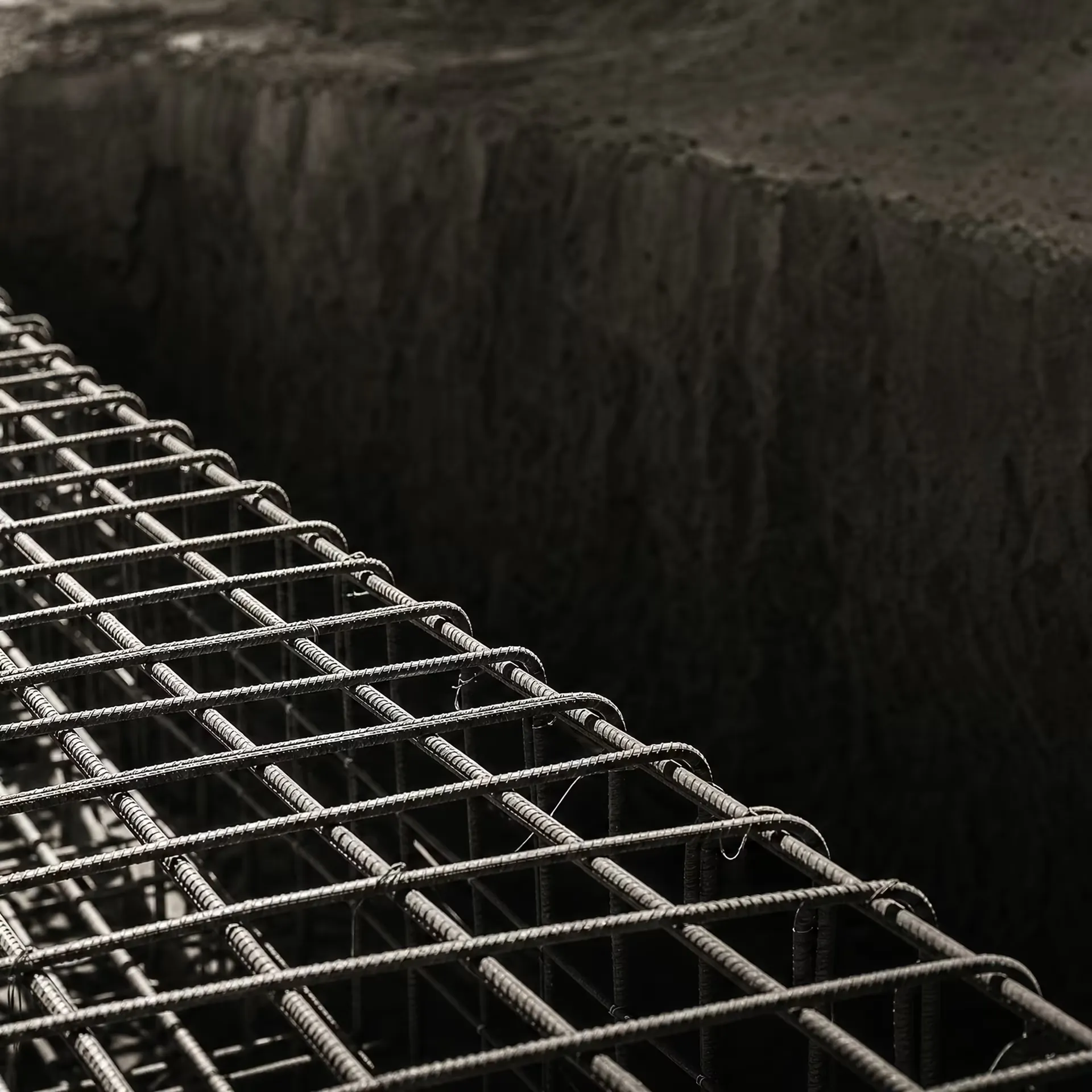 A dense orthogonal lattice of steel reinforcing bars tied into a concrete foundation trench in warm-tone monochrome, the ribbed bars receding under grazing light before the pour.