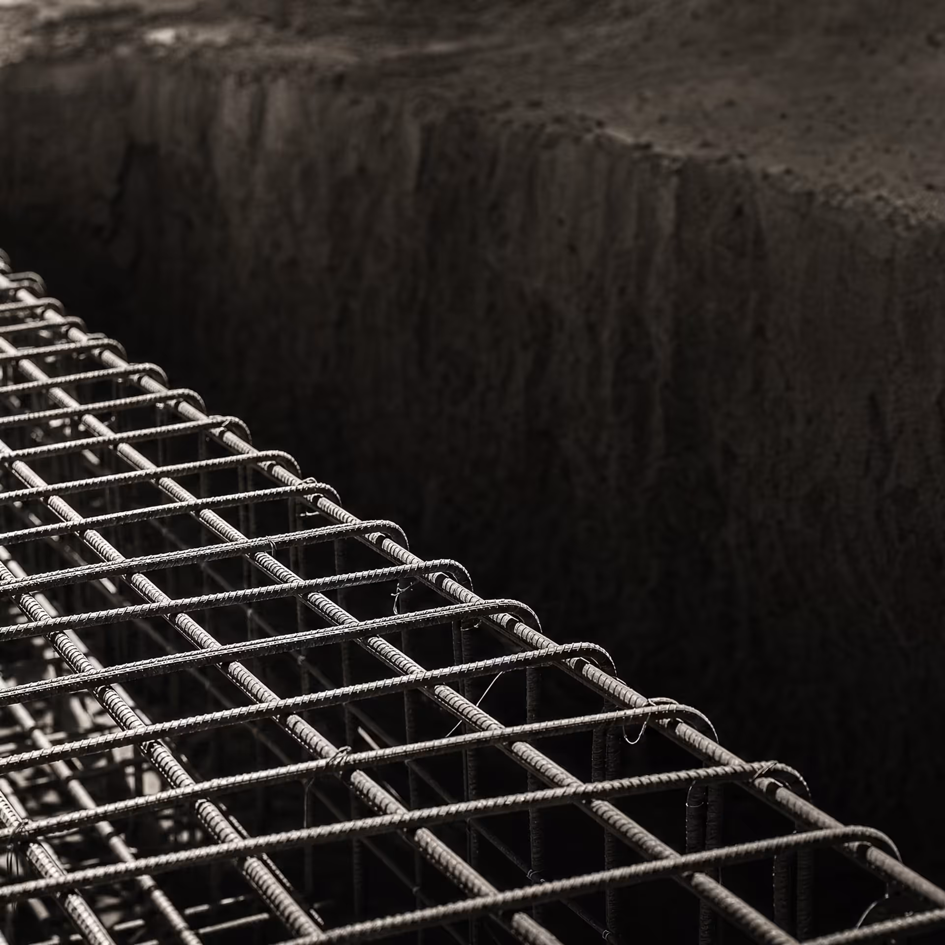 A dense orthogonal lattice of steel reinforcing bars tied into a concrete foundation trench in warm-tone monochrome, the ribbed bars receding under grazing light before the pour.