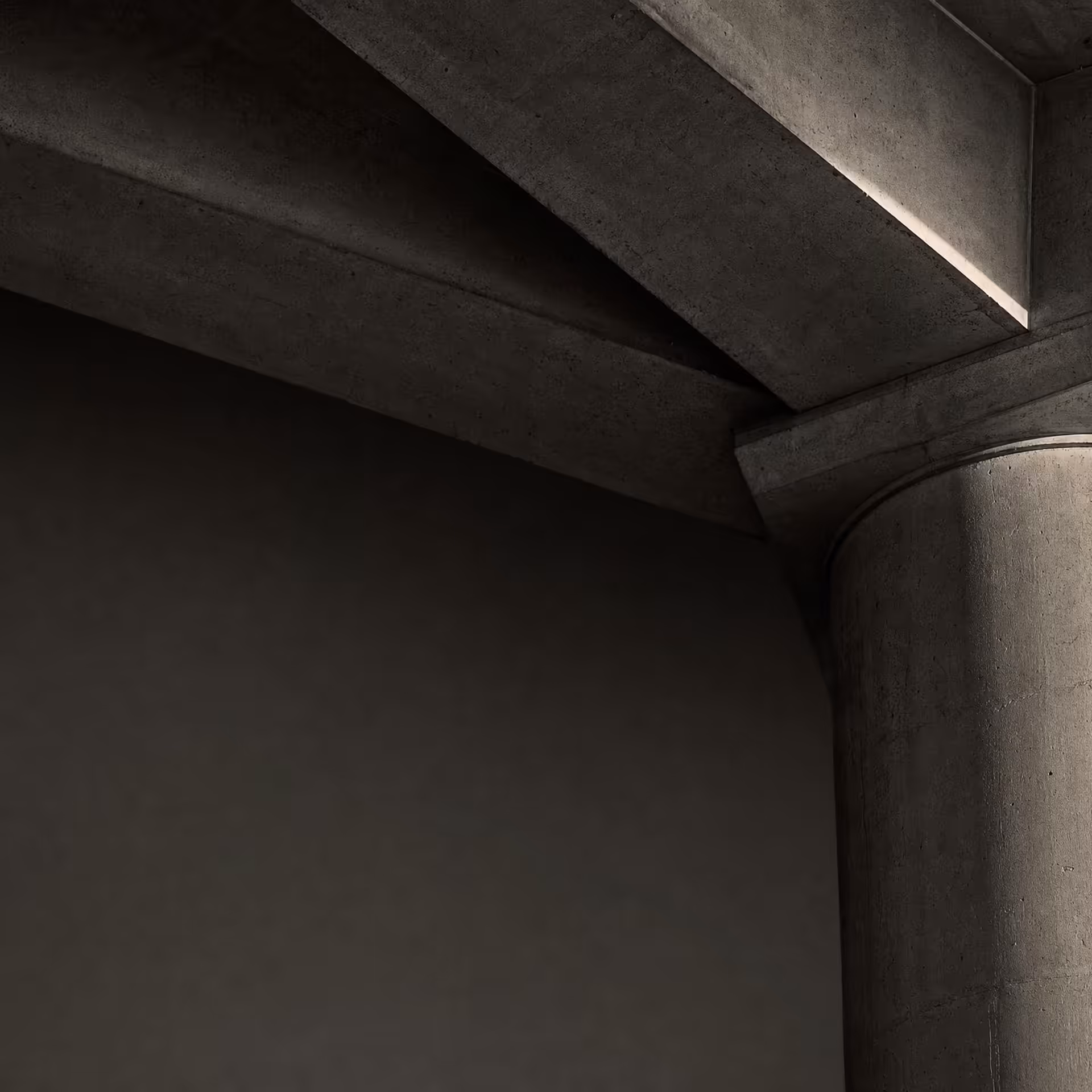 A single concrete column taking the load of three converging beams in warm-tone monochrome, the junction sharp under grazing daylight against receding tonal depth.