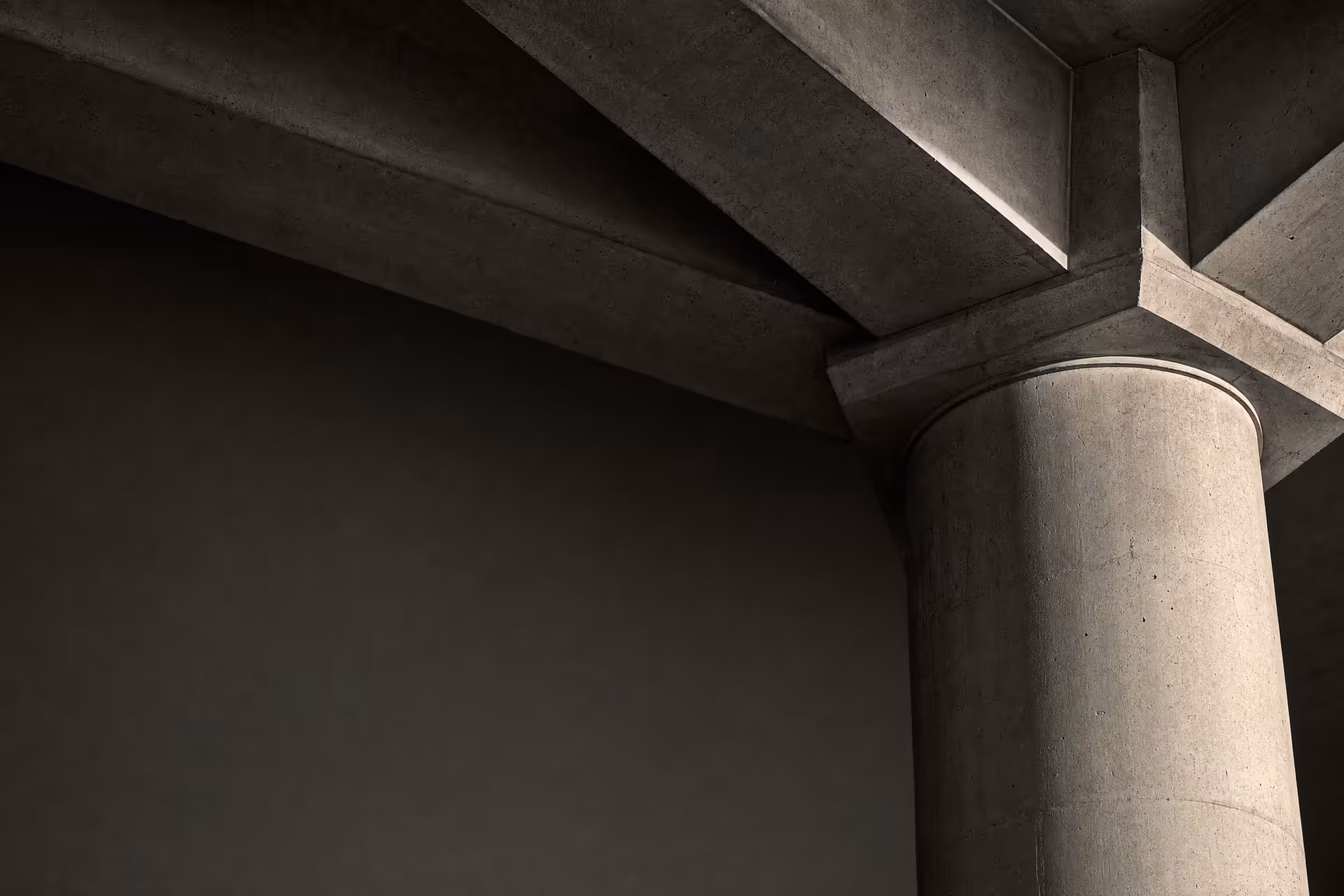 A single concrete column taking the load of three converging beams in warm-tone monochrome, the junction sharp under grazing daylight against receding tonal depth.