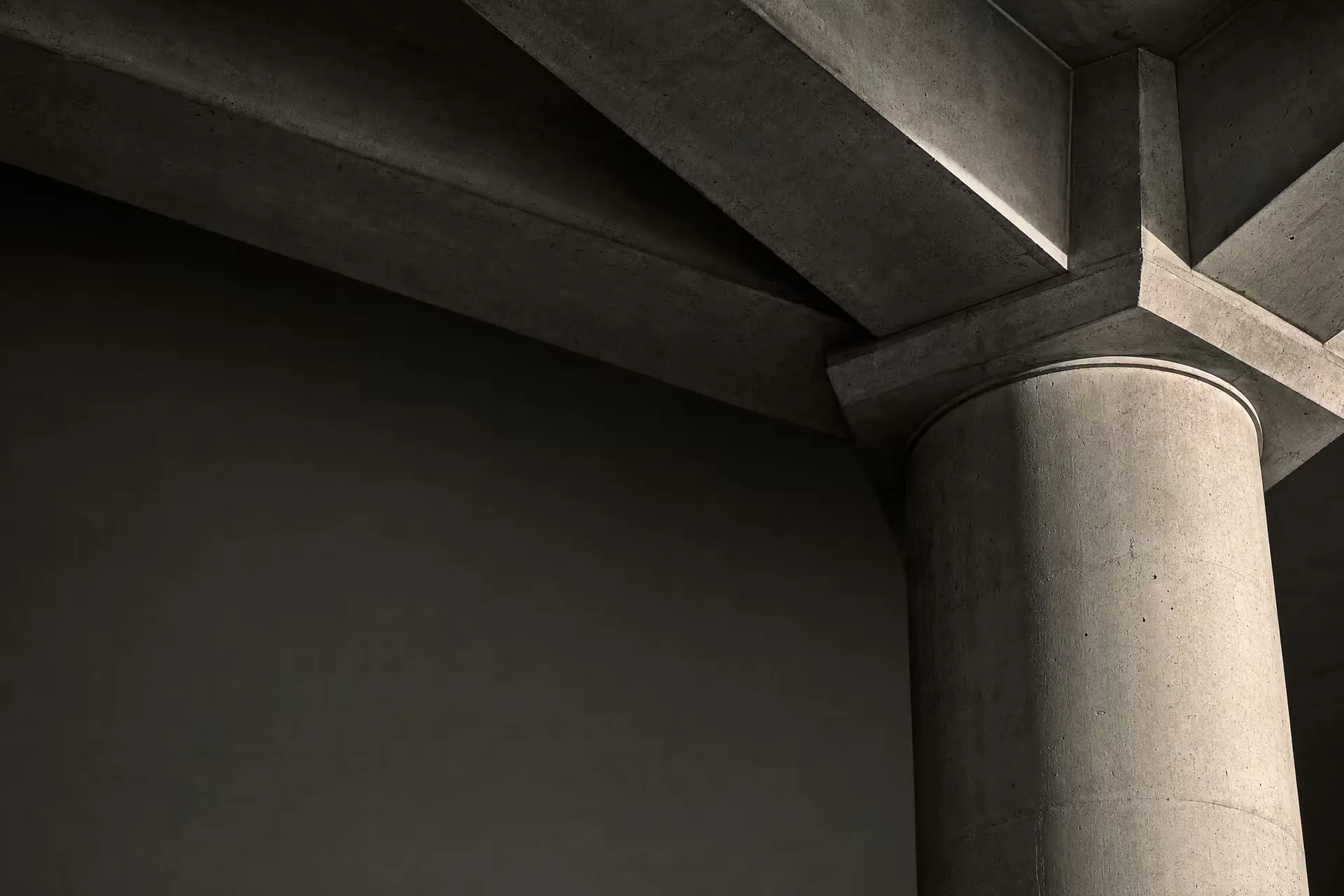 A single concrete column taking the load of three converging beams in warm-tone monochrome, the junction sharp under grazing daylight against receding tonal depth.