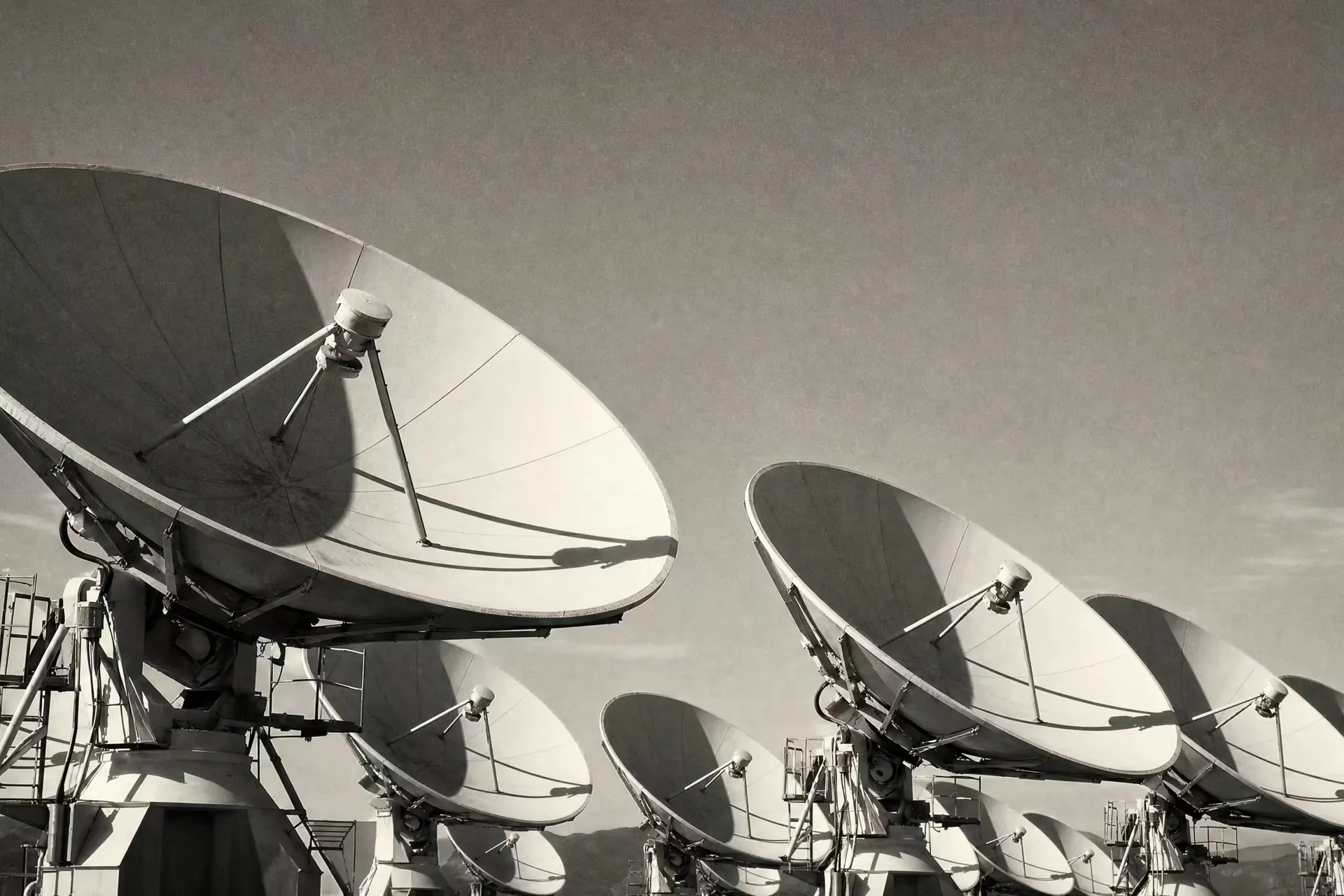 A satellite ground station, an array of large parabolic communication dishes angled skyward, warm-tone monochrome.