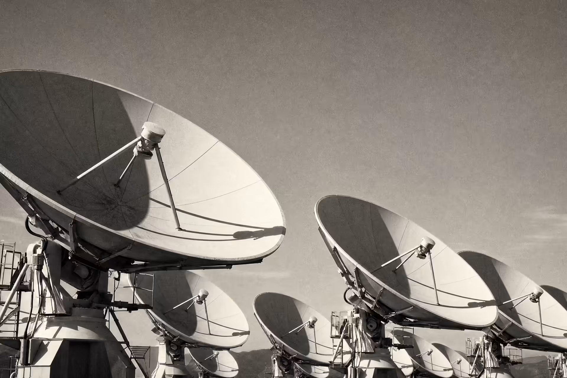 A satellite ground station, an array of large parabolic communication dishes angled skyward, warm-tone monochrome.