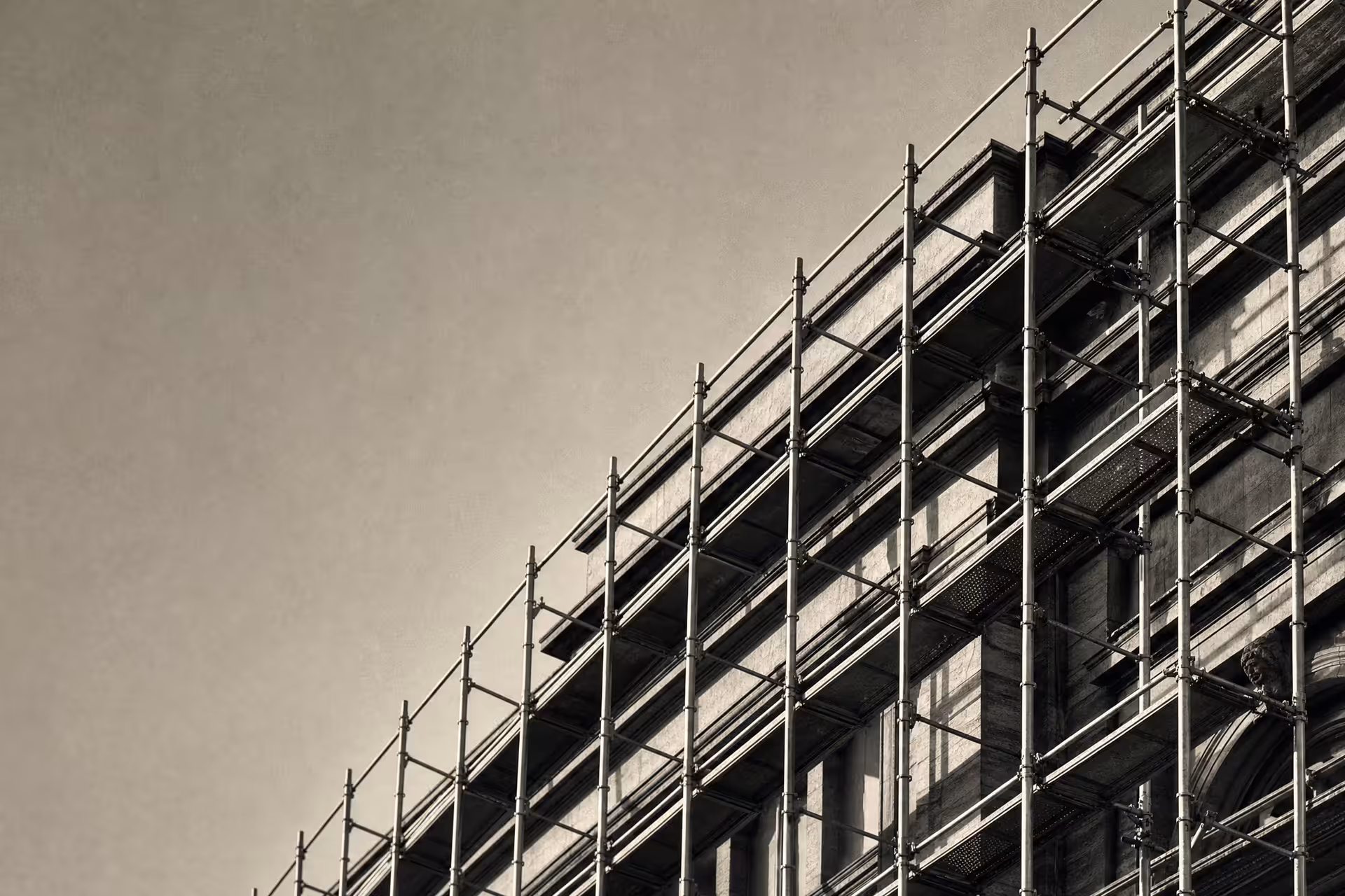 Steel scaffolding wrapping a stone building mid-renovation in warm-tone monochrome, the new tubular frame standing in front of the older facade as distinct planes.