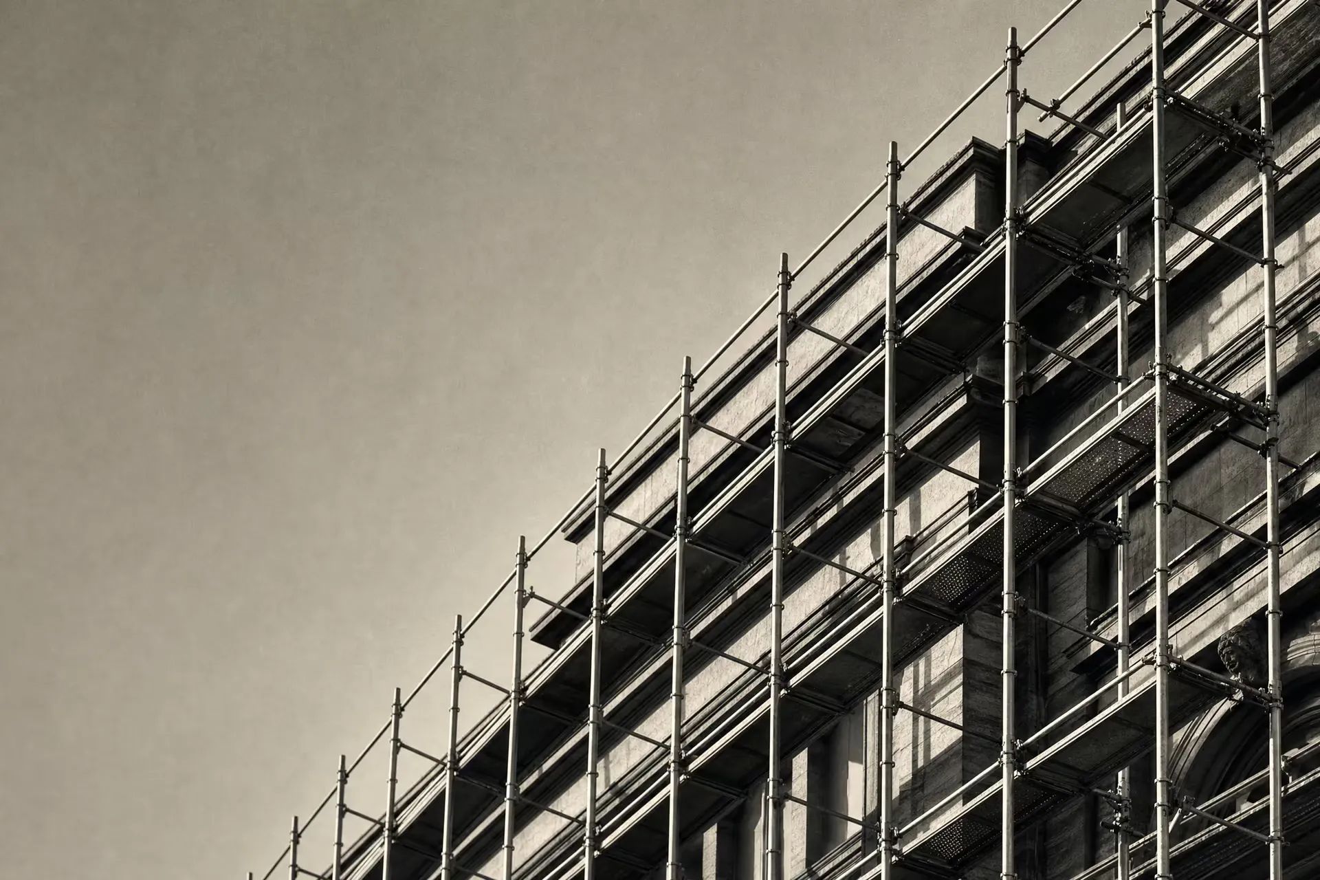 Steel scaffolding wrapping a stone building mid-renovation in warm-tone monochrome, the new tubular frame standing in front of the older facade as distinct planes.