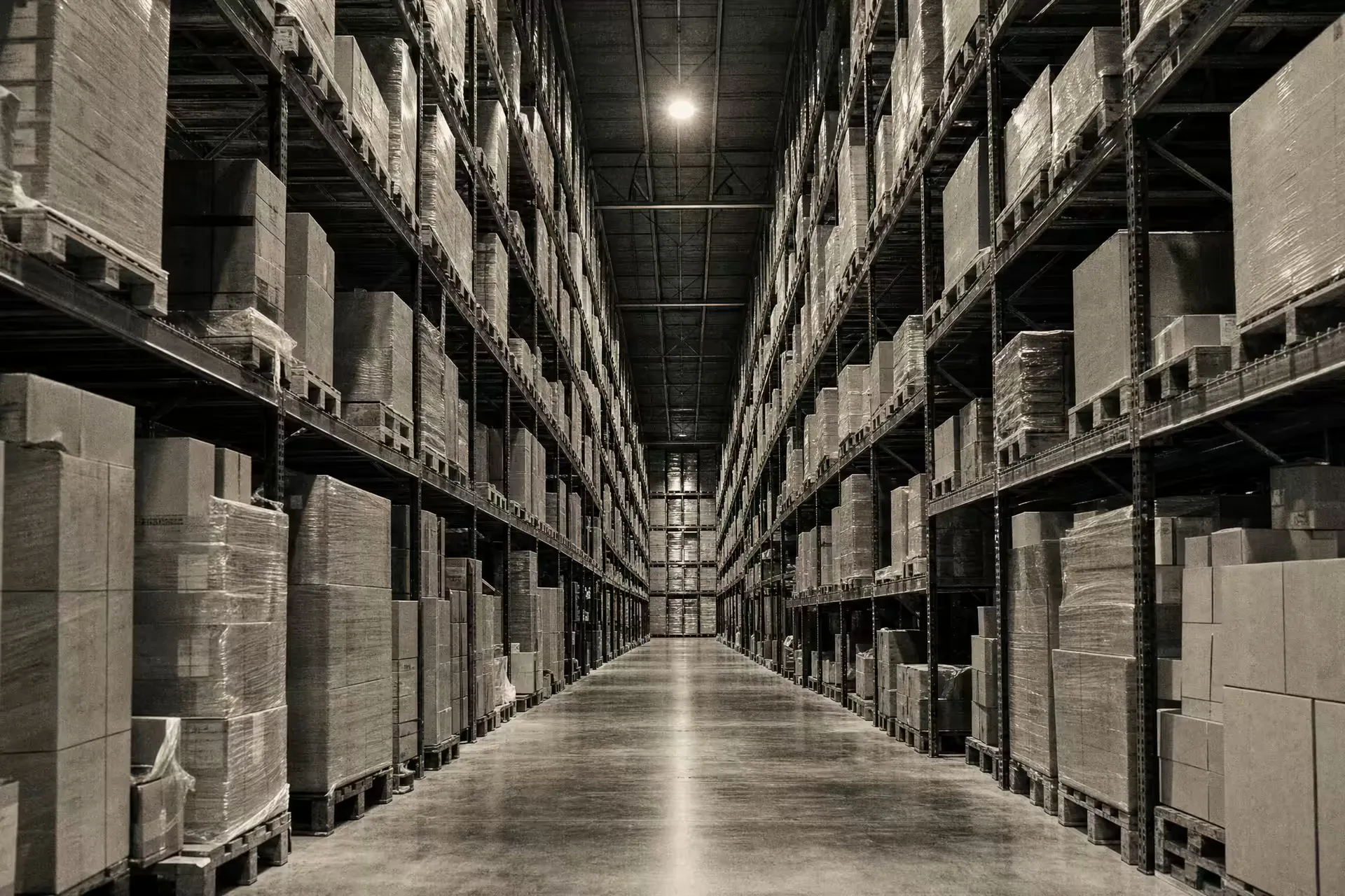 A stocked logistics warehouse, tall pallet racking receding down a long aisle, warm-tone monochrome.