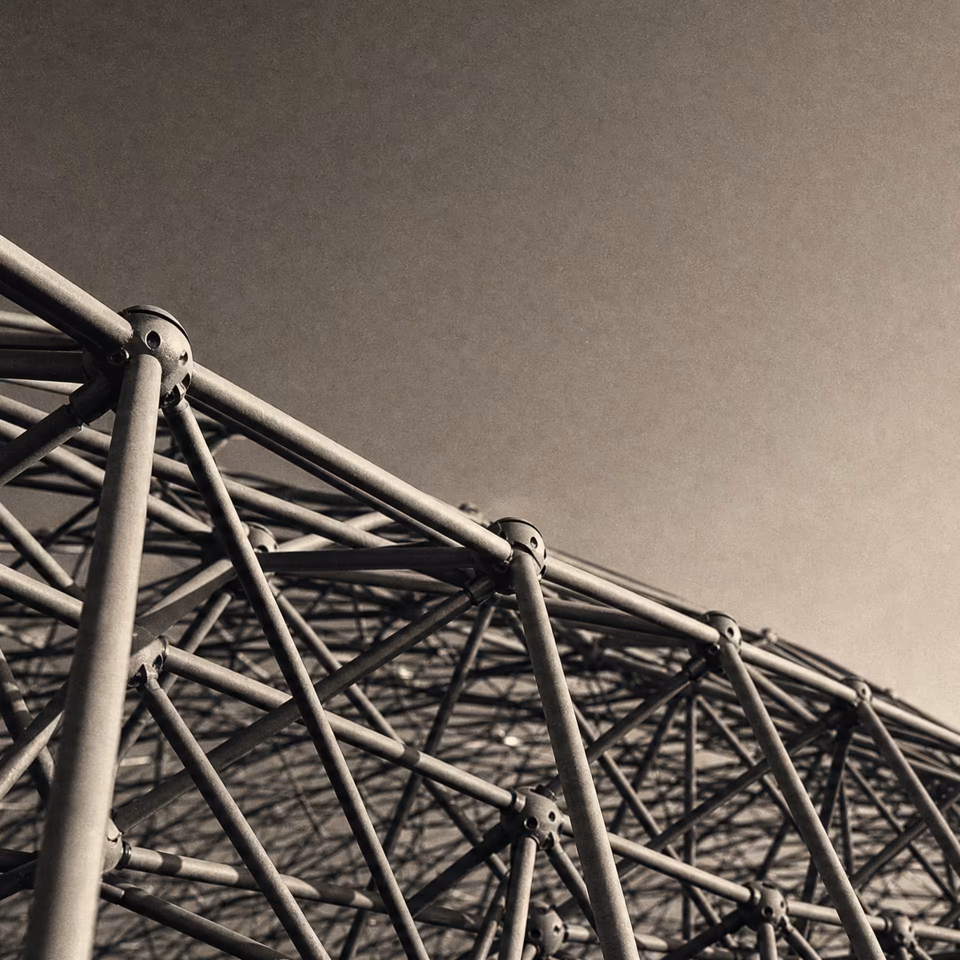 A steel space-frame truss seen from below in warm-tone monochrome, triangulated members receding to a vanishing point against open sky.