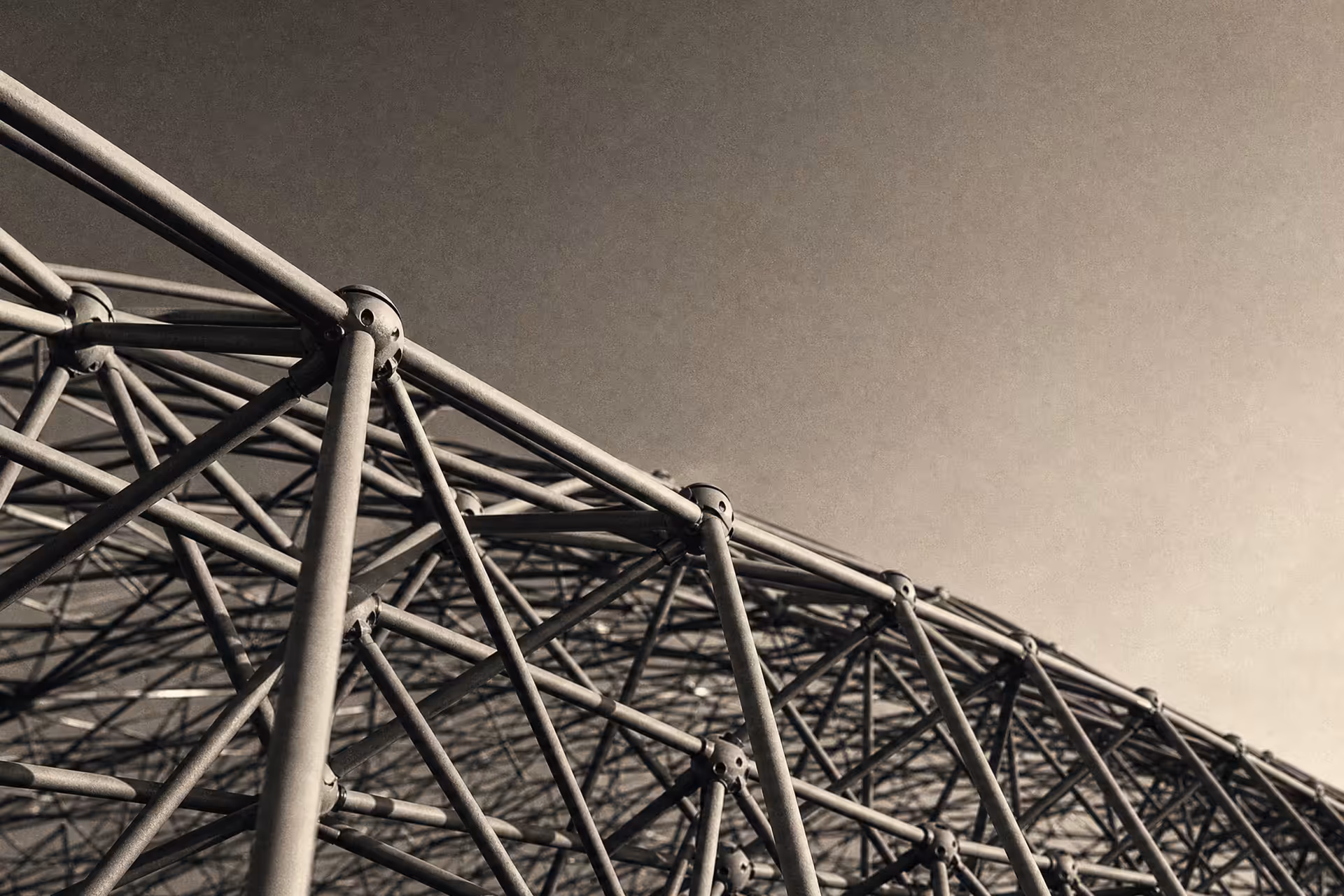 A steel space-frame truss seen from below in warm-tone monochrome, triangulated members receding to a vanishing point against open sky.