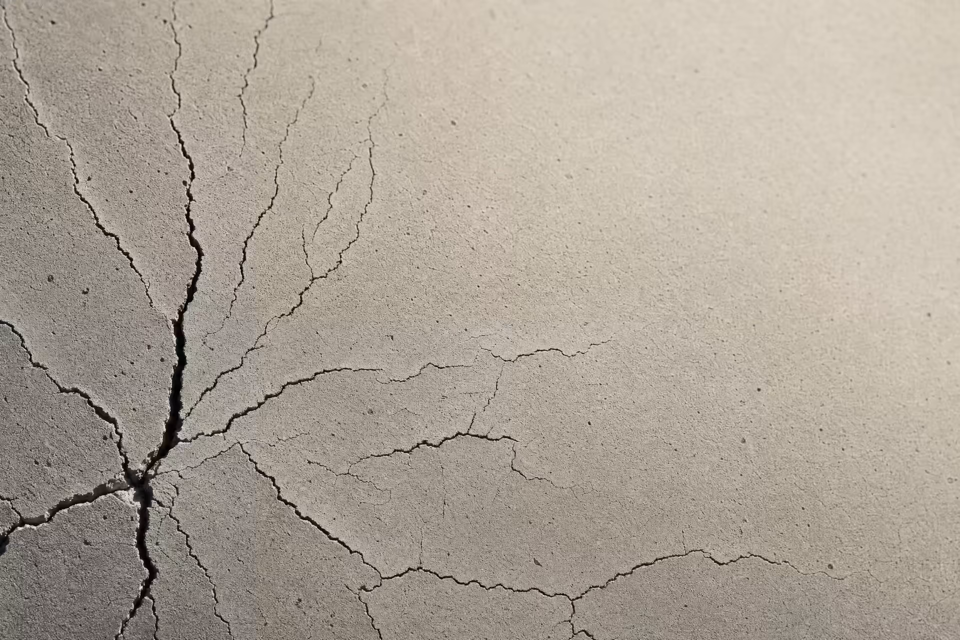 Fine fatigue cracks branching from a single point across a concrete surface in warm-tone monochrome, the fissures thrown into relief by grazing light.