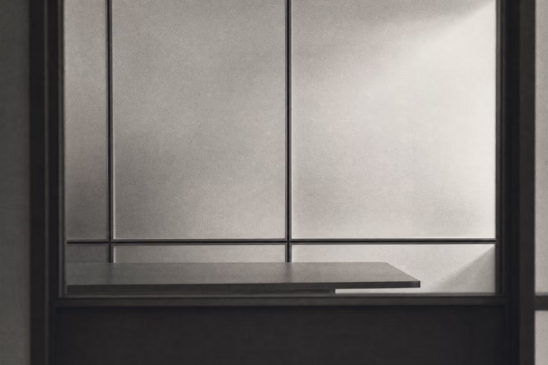 An empty meeting room seen through a frosted-glass partition, hairline aluminium frames dividing the panels into rectilinear sections, warm-tone monochrome.