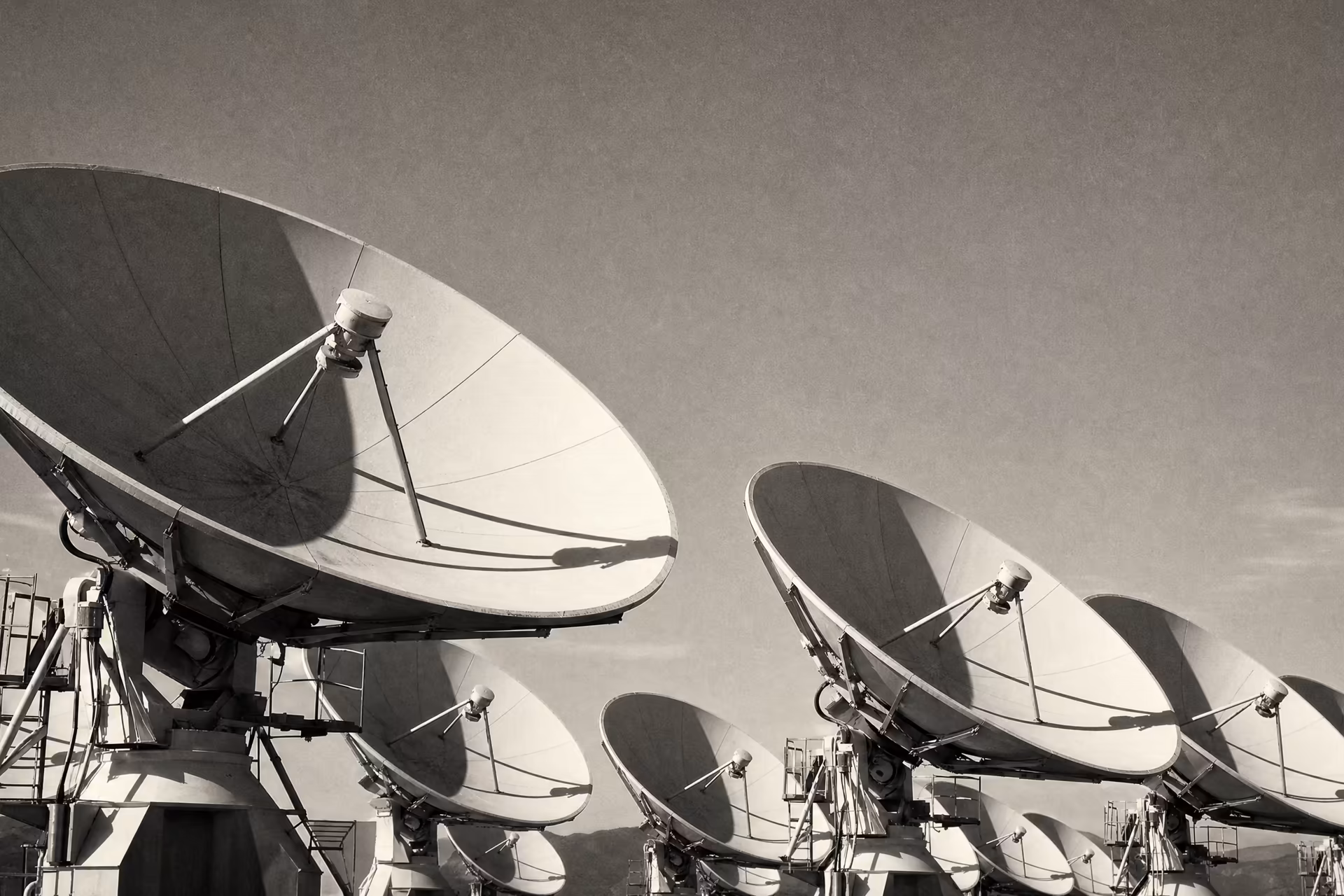 A satellite ground station, an array of large parabolic communication dishes angled skyward, warm-tone monochrome.