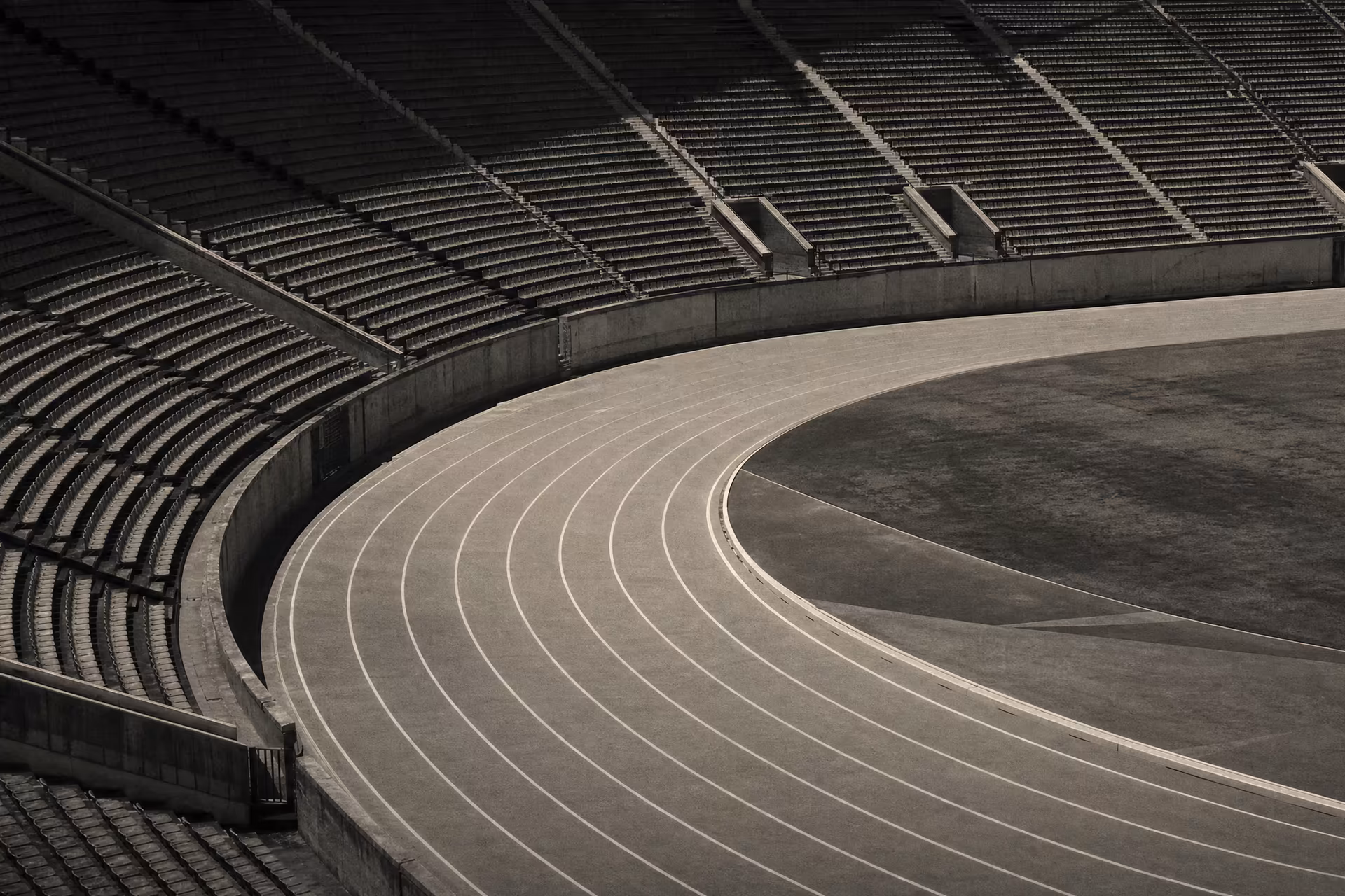 An athletics stadium running track, curved sprint lanes sweeping through the bend below empty grandstands, warm-tone monochrome.